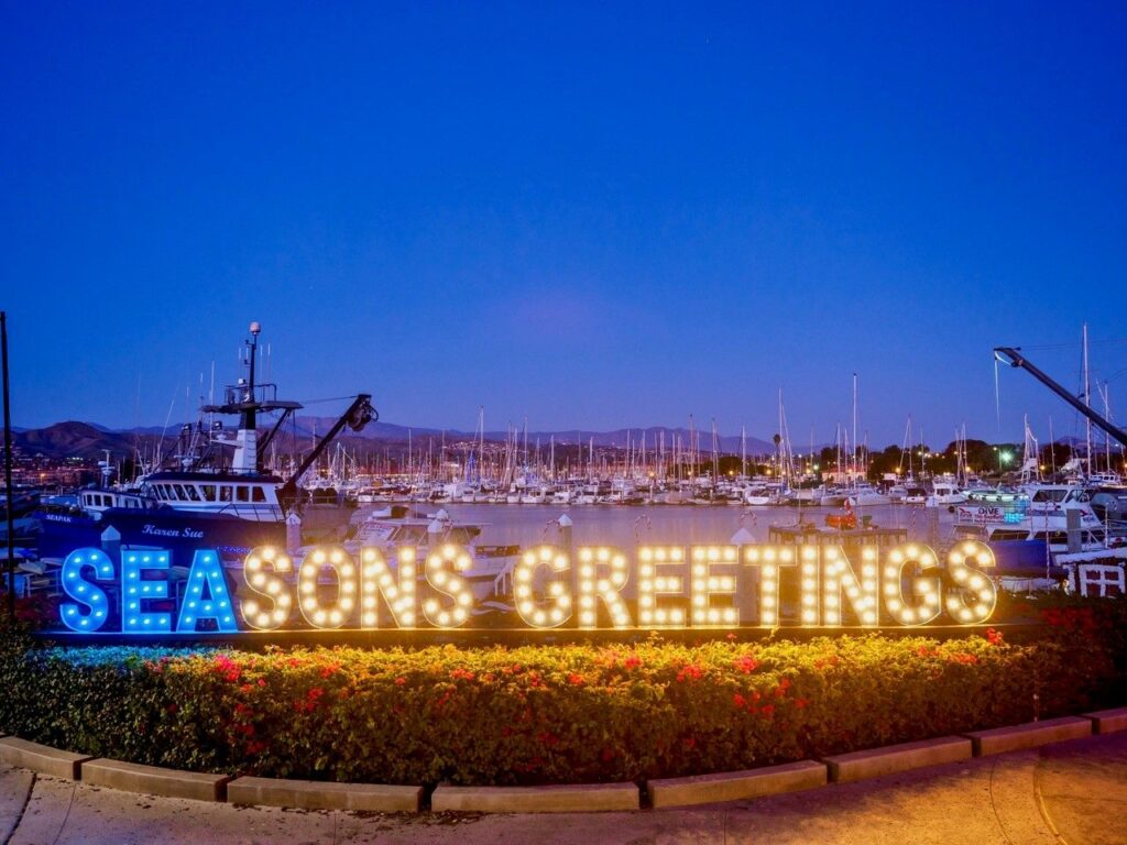 AQ lit up sign that says seasons greetings in blue and white with harbor marina the background