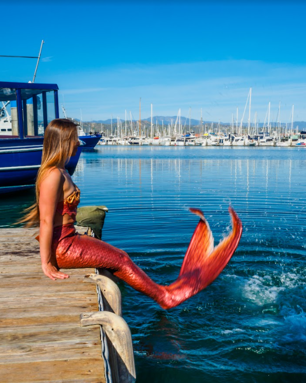 Mermaid with orange tail sitting on a dock in Ventura Harbor and splashing in the water
