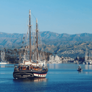 A large sailboat named "Mystic Whaler" in a harbor with mountain range and numerous sailboats in the background on a clear, sunny day.