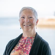 An older woman with short gray hair smiles while wearing a colorful patterned top, black sweater, and star-shaped earrings. She stands in front of an out-of-focus seaside background.