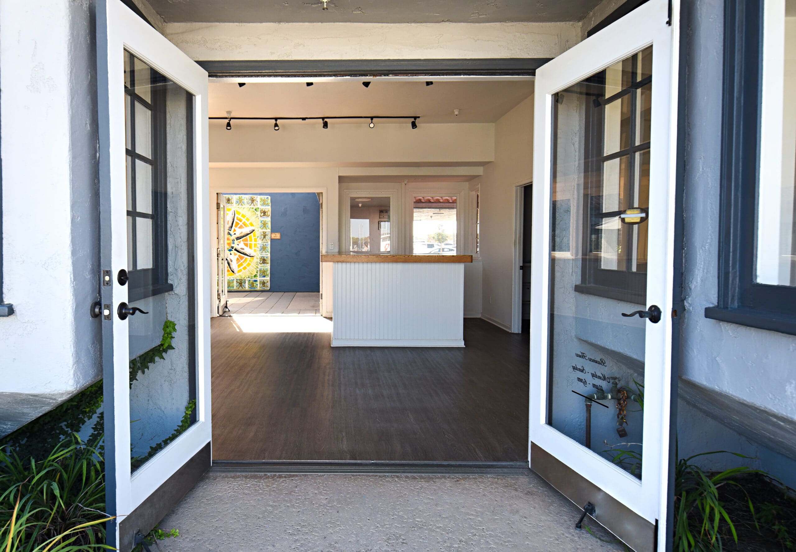 Open double doors lead into a bright room with wooden flooring, a white reception desk, and a colorful mosaic wall in the background.