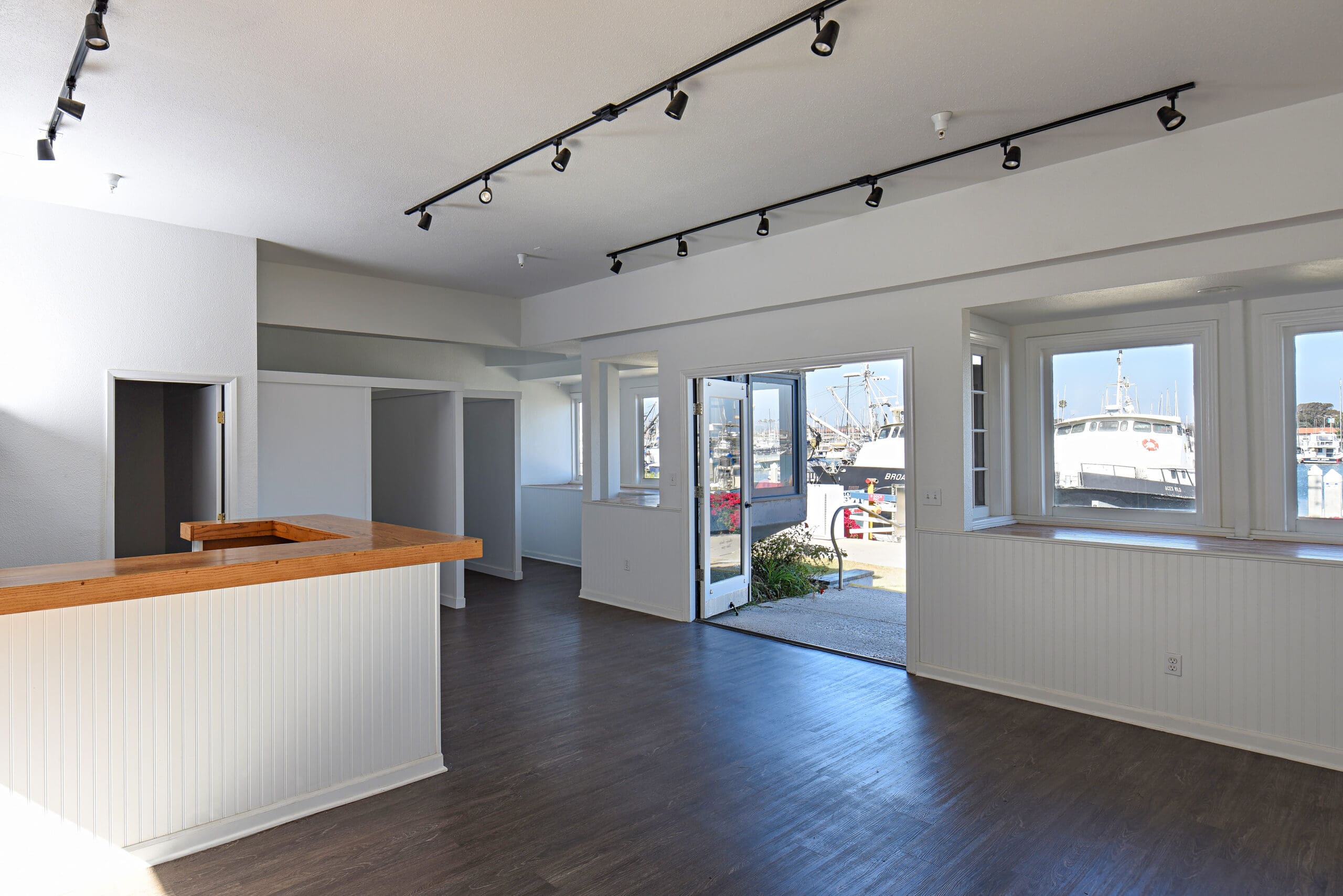 Empty commercial space with wooden counter, track lighting, and large windows showing boats outside. White walls and dark wood flooring create a clean, open atmosphere.