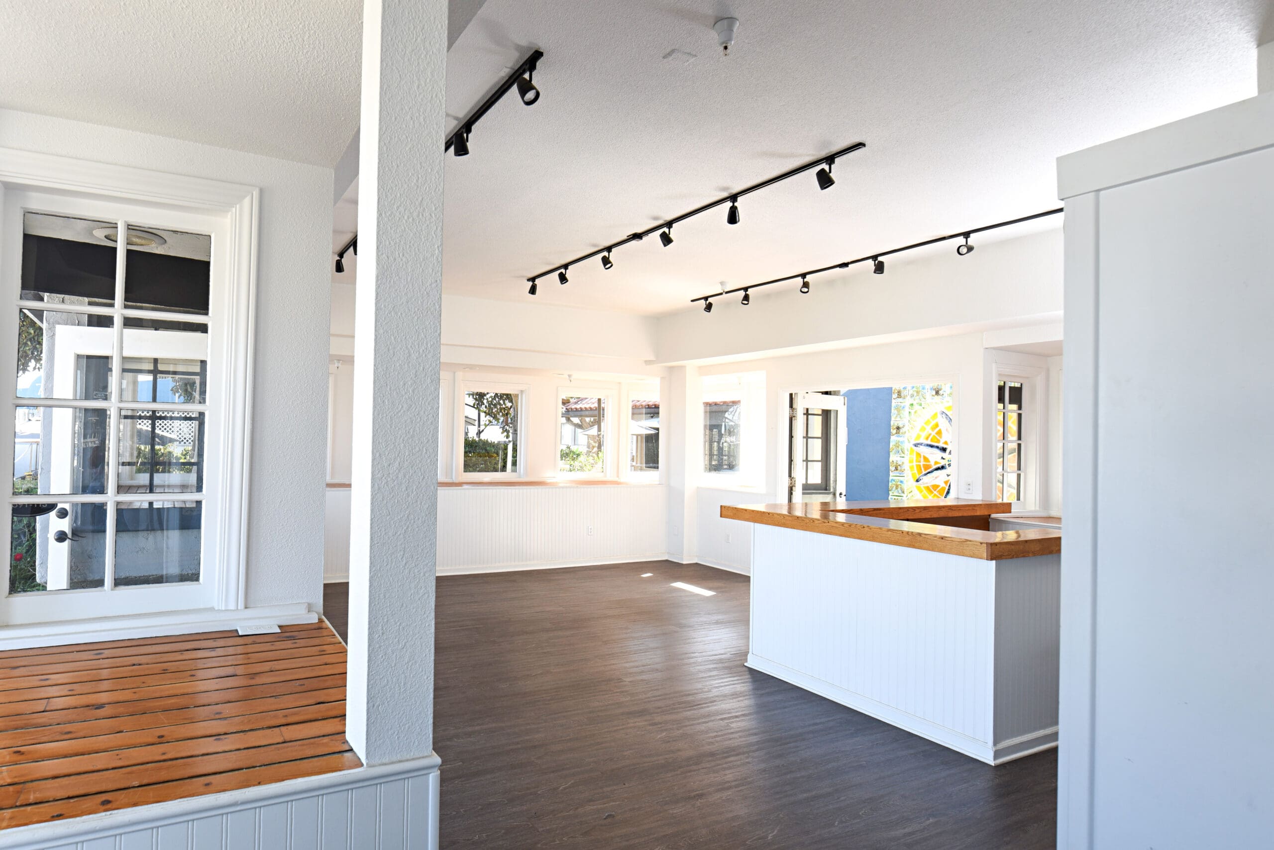 Interior of an empty, well-lit room featuring white walls, large windows, a wooden counter, and track lighting on the ceiling.