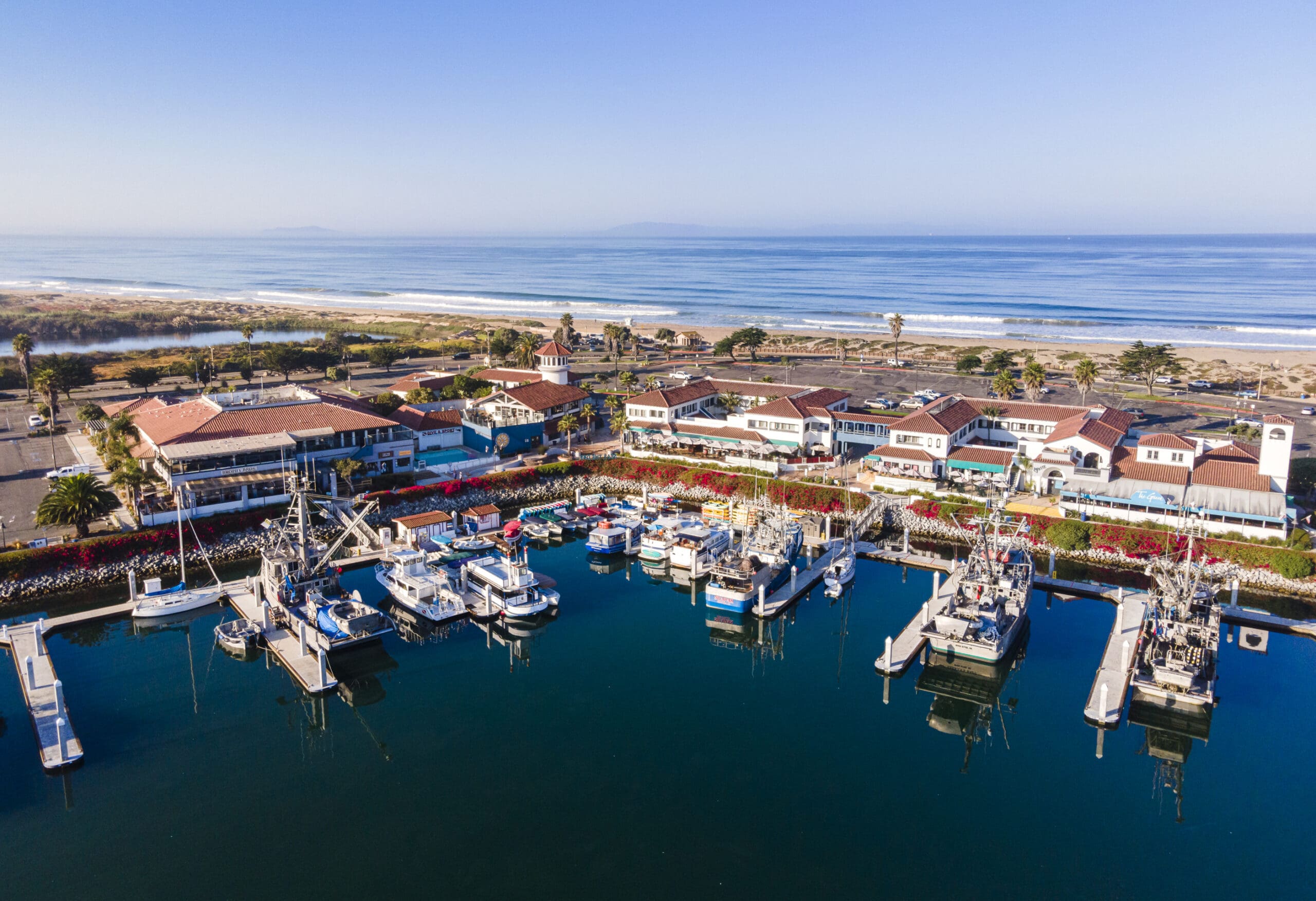 Aerial view of a marina with docked boats, adjacent buildings, and a coastal beach in the background under a clear blue sky.