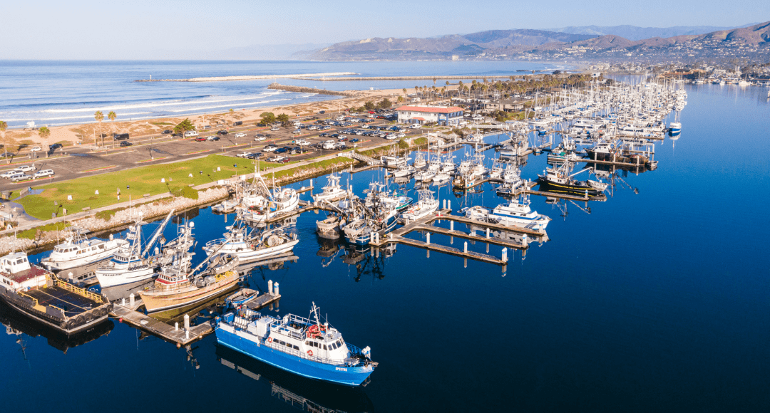 Aerial view of a coastal marina with numerous boats docked in clear blue water. The surrounding area includes parking, green spaces, and a distant hilly landscape.