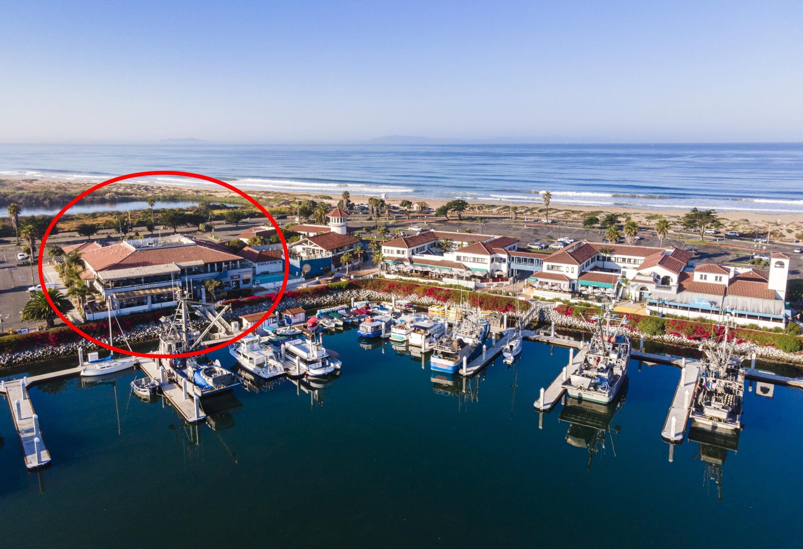 Aerial view of a marina with several boats docked. A group of buildings and a red circle highlight a specific area near the shoreline.