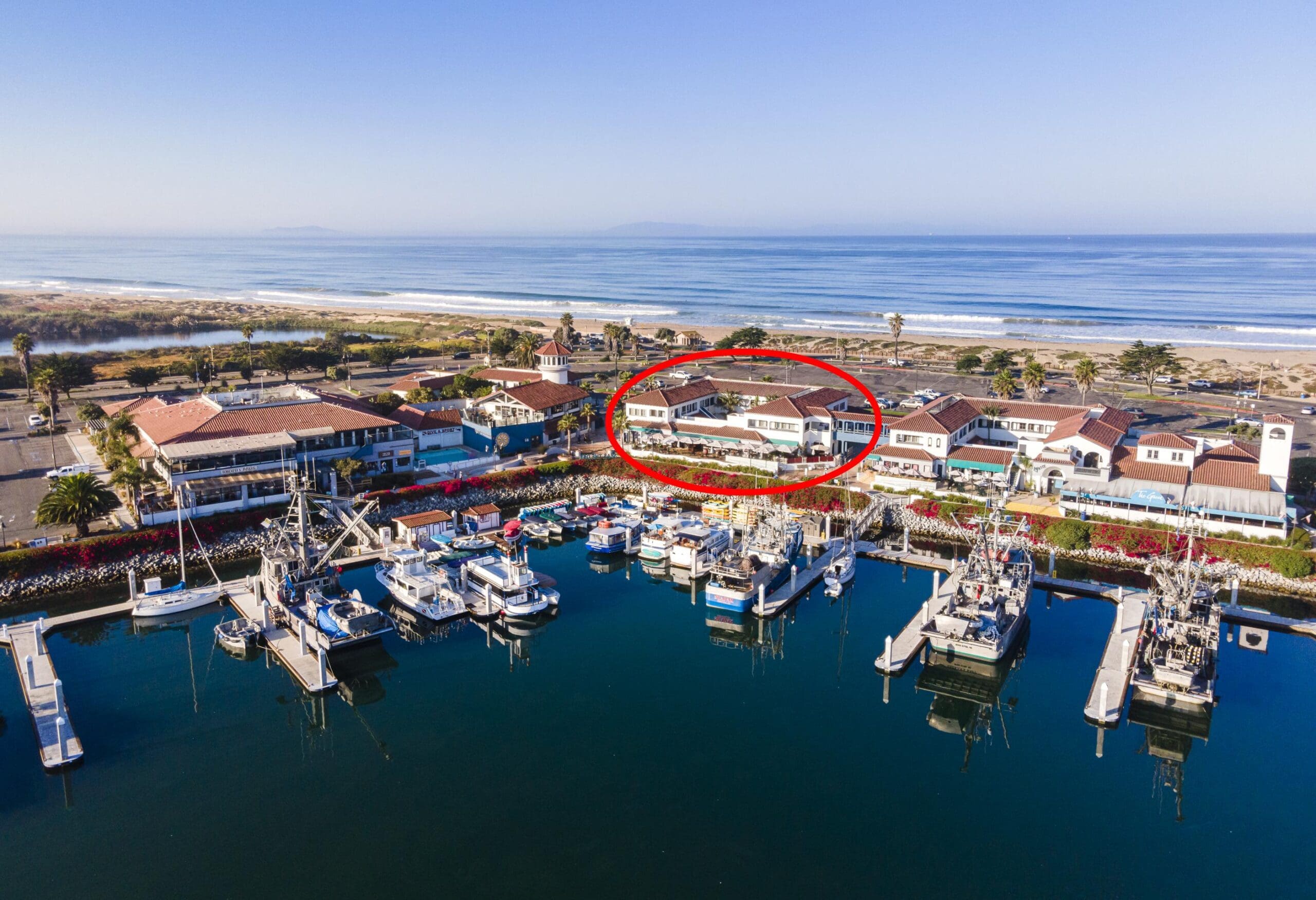 Aerial view of a marina with parked boats and surrounding buildings near the beach. One building is circled in red.