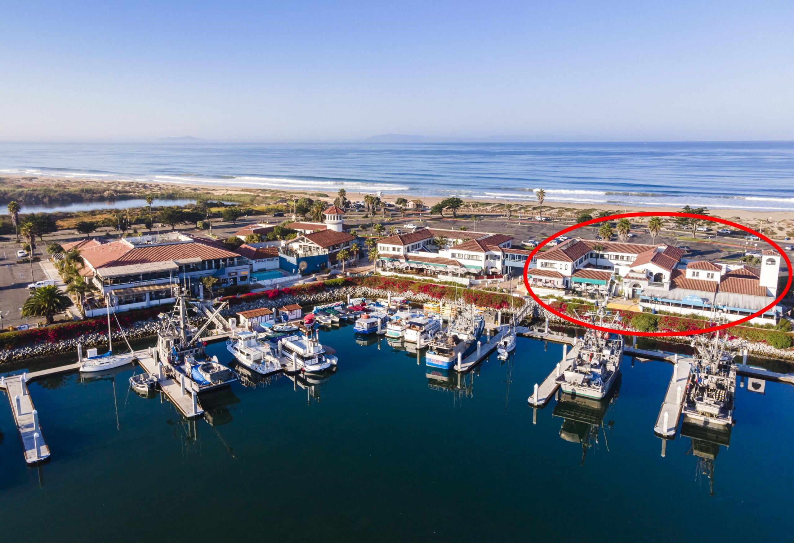 Aerial view of a marina with multiple boats docked. Nearby, there are several buildings; one is circled in red. The ocean and beach are visible in the background.