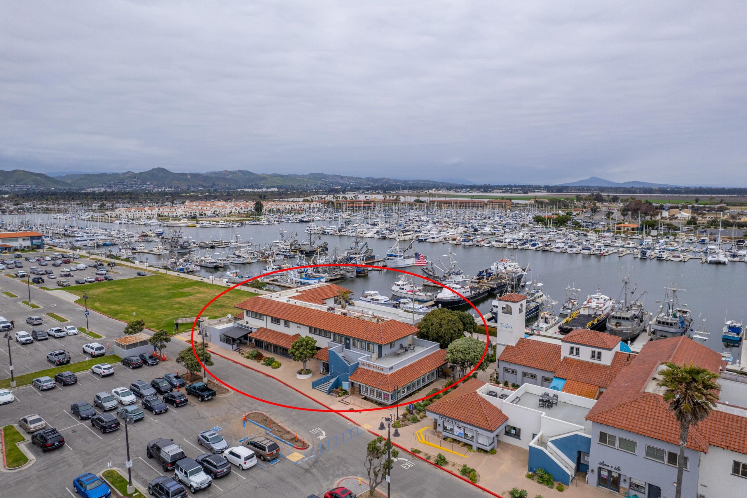 Aerial view of a marina with numerous boats docked. A building with a red roof is circled, situated near a parking lot and a grassy area. Overcast sky in the background.