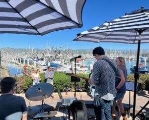 A small band performs outdoors near a marina with boats and mountains in the background; striped umbrellas provide shade.