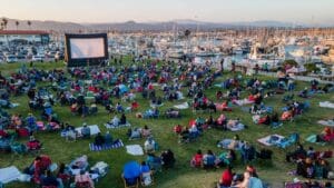 Large crowd seated on a grassy area watching an outdoor movie on a big screen near a marina filled with boats, during daylight.