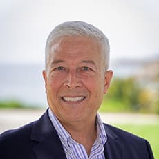 A man with short gray hair wearing a navy blazer and striped shirt is smiling outdoors, with greenery and water blurred in the background.