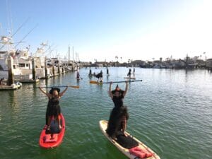 People dressed as witches paddle on stand-up paddleboards and kayaks in a marina, with boats and other participants visible in the background.